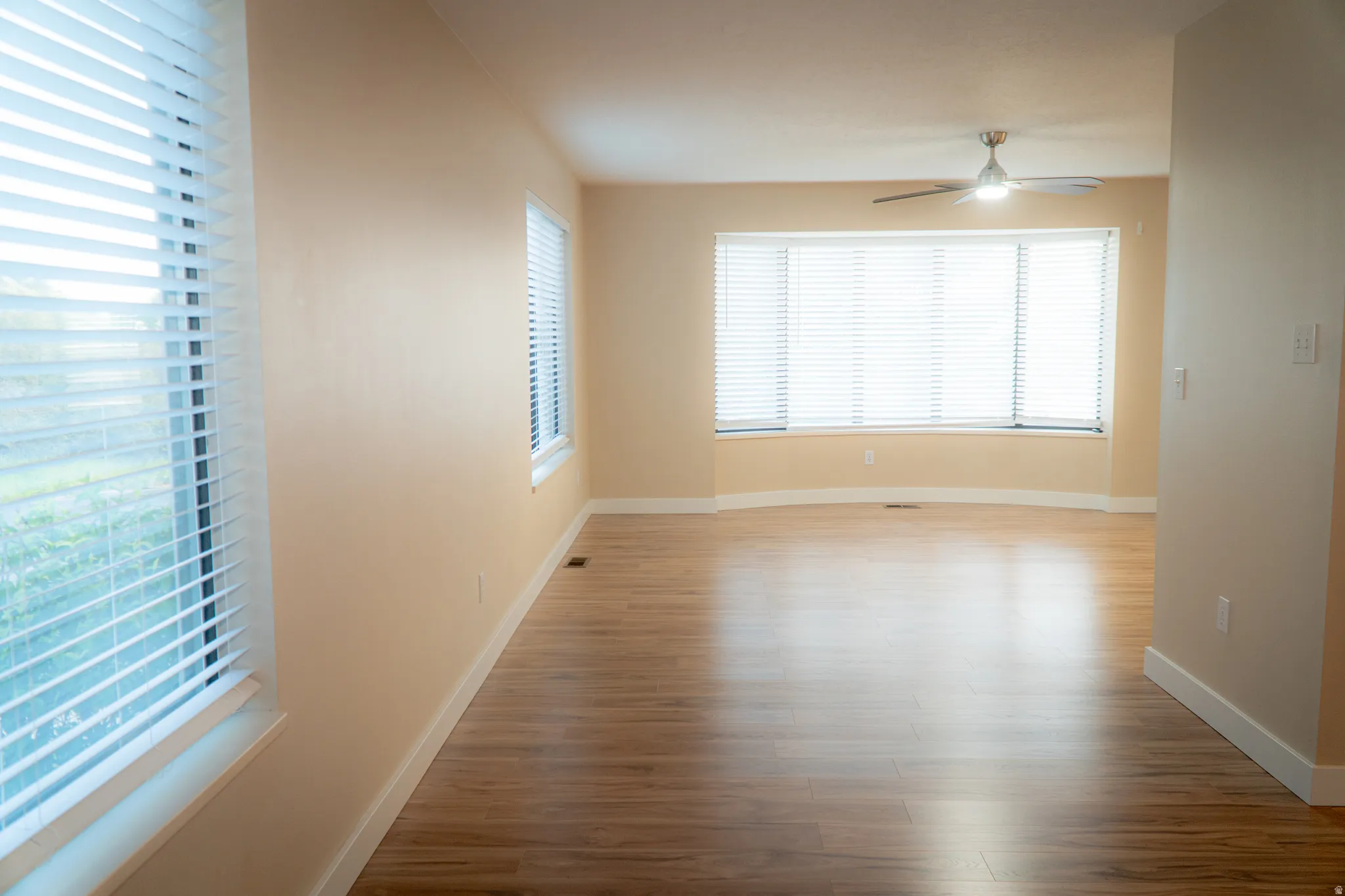 Spacious room featuring wood-finish flooring, multiple windows with blinds, a large bay window, and a ceiling fan with integrated lighting
