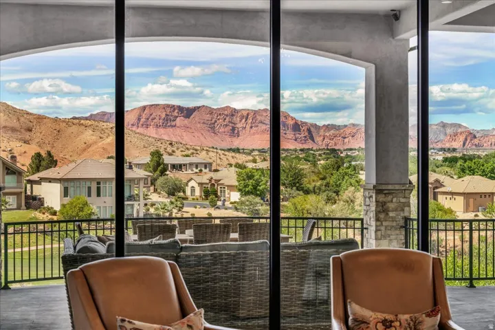 Expansive sliding glass doors open to an outdoor living area with a stone pillar and black metal railing, featuring distant red rock formations and verdant landscape