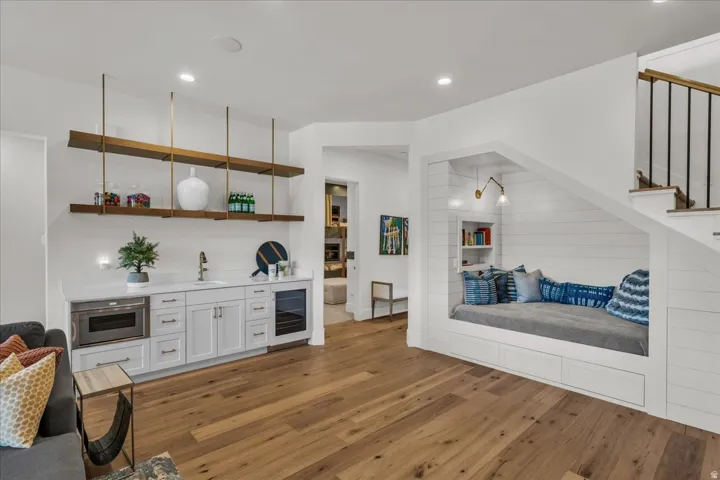 Hardwood flooring throughout a versatile space featuring a built-in wet bar with under-counter microwave and beverage cooler, and a custom window seat with integrated shelving and wall sconce lighting