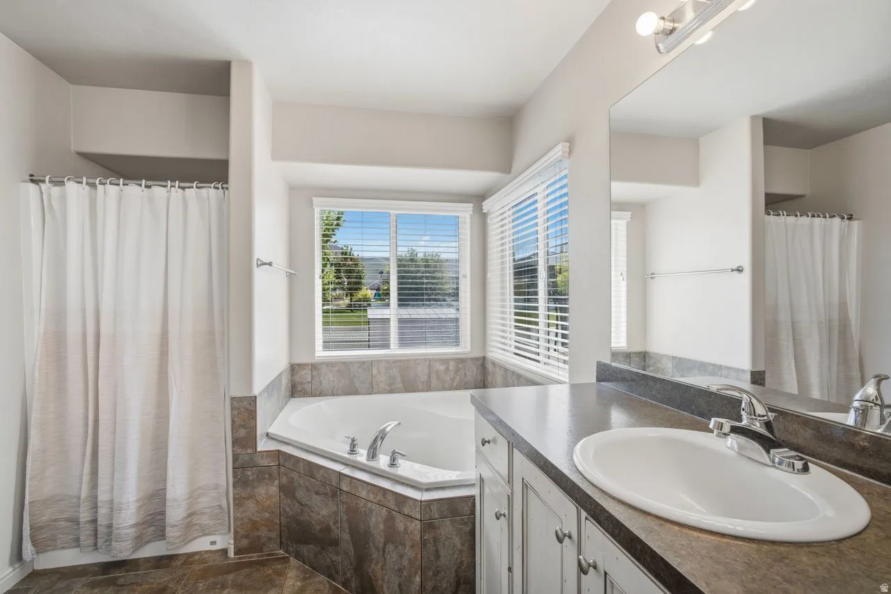 Bathroom featuring a corner soaking tub with surrounding tile, a window with blinds, and a vanity with an integrated sink and mirror