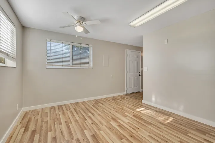 Spacious room featuring wood-finish flooring, neutral-toned walls, multiple windows with blinds, a ceiling fan with integrated lighting, and a white paneled door