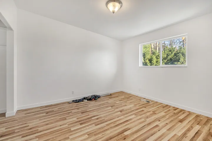 Bright room featuring wide plank wood-finish flooring, white baseboards, a flush-mount ceiling light fixture, and a large window providing a verdant outdoor scene