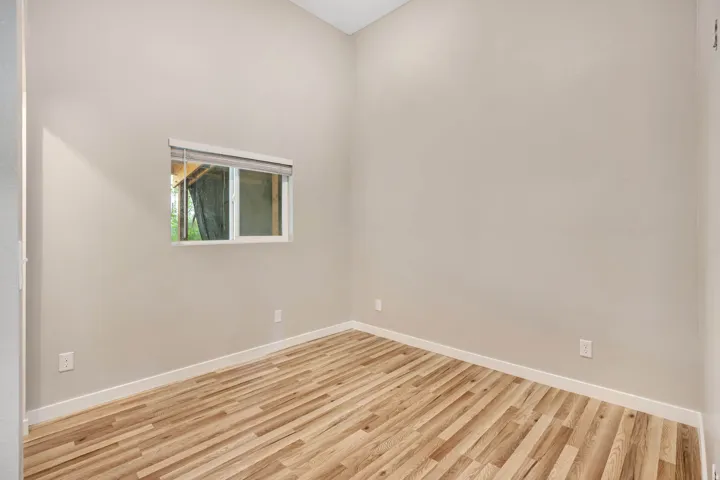 Vaulted ceiling room featuring wood-finish flooring, a white trim window with roller shade, and neutral wall paint