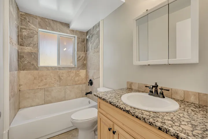 Bathroom featuring a white tub, natural stone-look wall tiling, a light wood-finish vanity with an integrated sink, a granite-look countertop, and a mirrored medicine cabinet