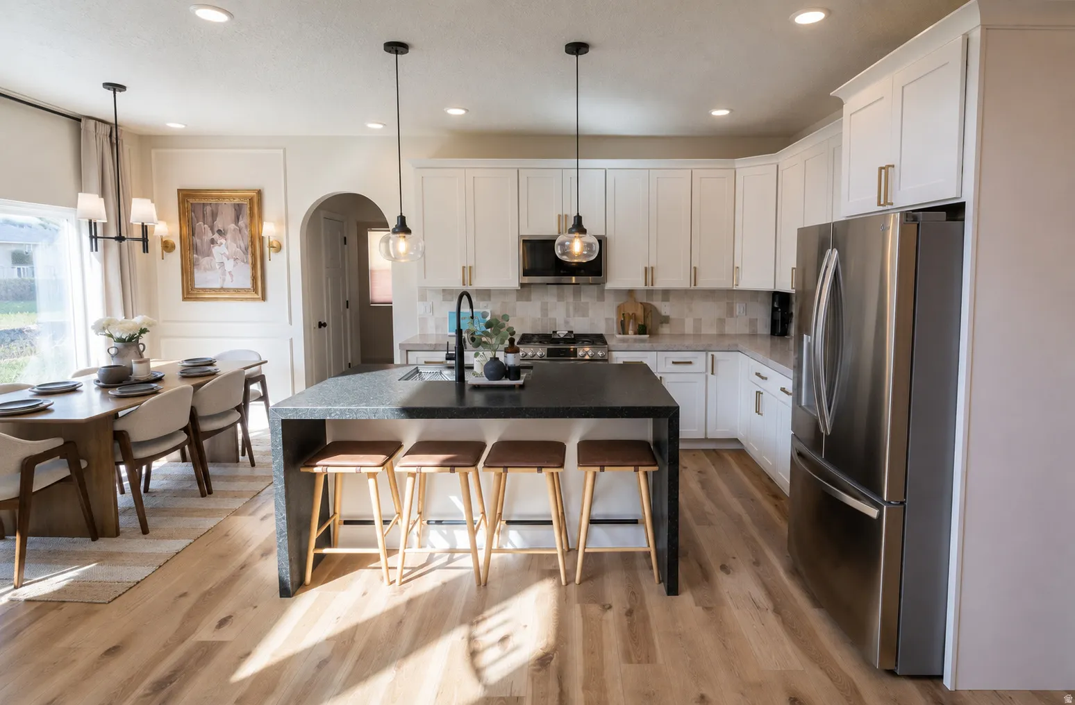 Open-concept kitchen and dining area featuring light wood-finish flooring, white cabinetry with gold-tone hardware, a dark-toned island with integrated seating, and stainless steel appliances