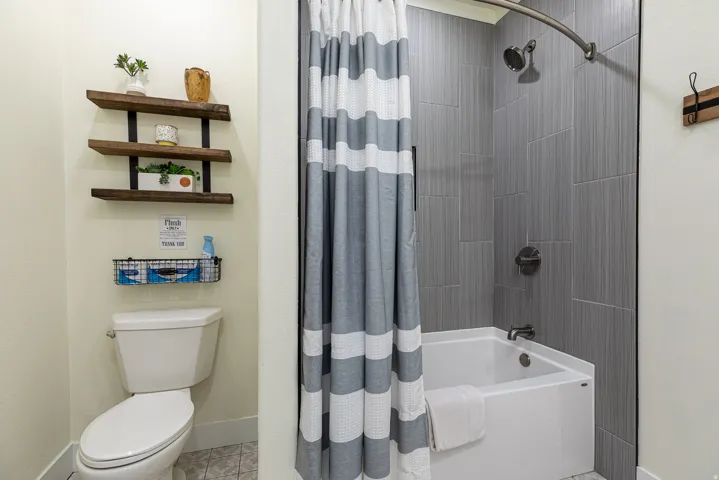 Bathroom featuring a shower-tub combination with gray vertical tile surround, a white porcelain toilet, and three-tier wall-mounted shelving above
