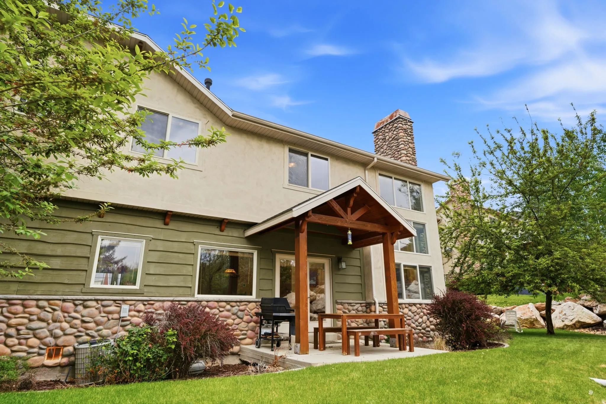Rear exterior featuring a stone chimney, covered patio with wood beam accents, multiple windows, and a stone foundation