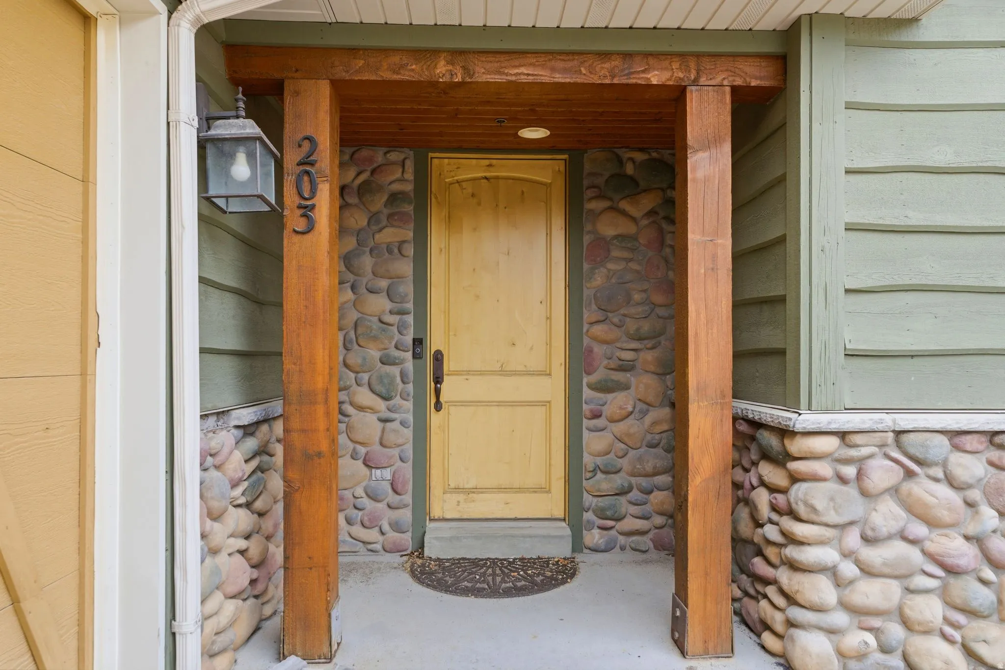Entryway featuring a natural wood door, stacked stone facade, exposed wood beams, and a concrete porch