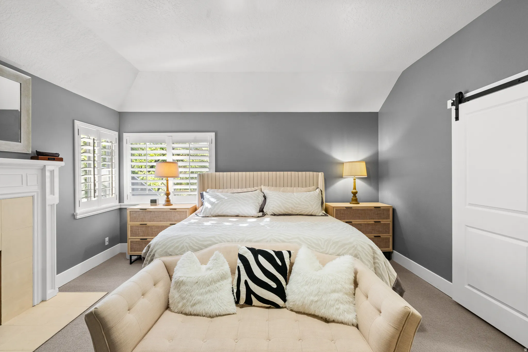 Vaulted ceiling room featuring a decorative fireplace with white mantel, two windows with plantation shutters, a sliding barn door, and light-toned carpet flooring