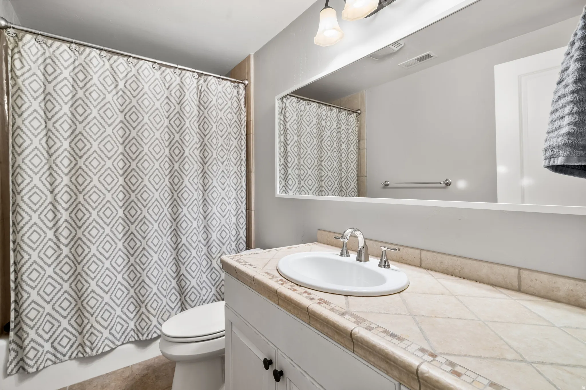 Bathroom featuring a tiled vanity countertop with an undermount sink, white vanity cabinetry with dark hardware, a large wall-mounted mirror, and a wall-mounted light fixture