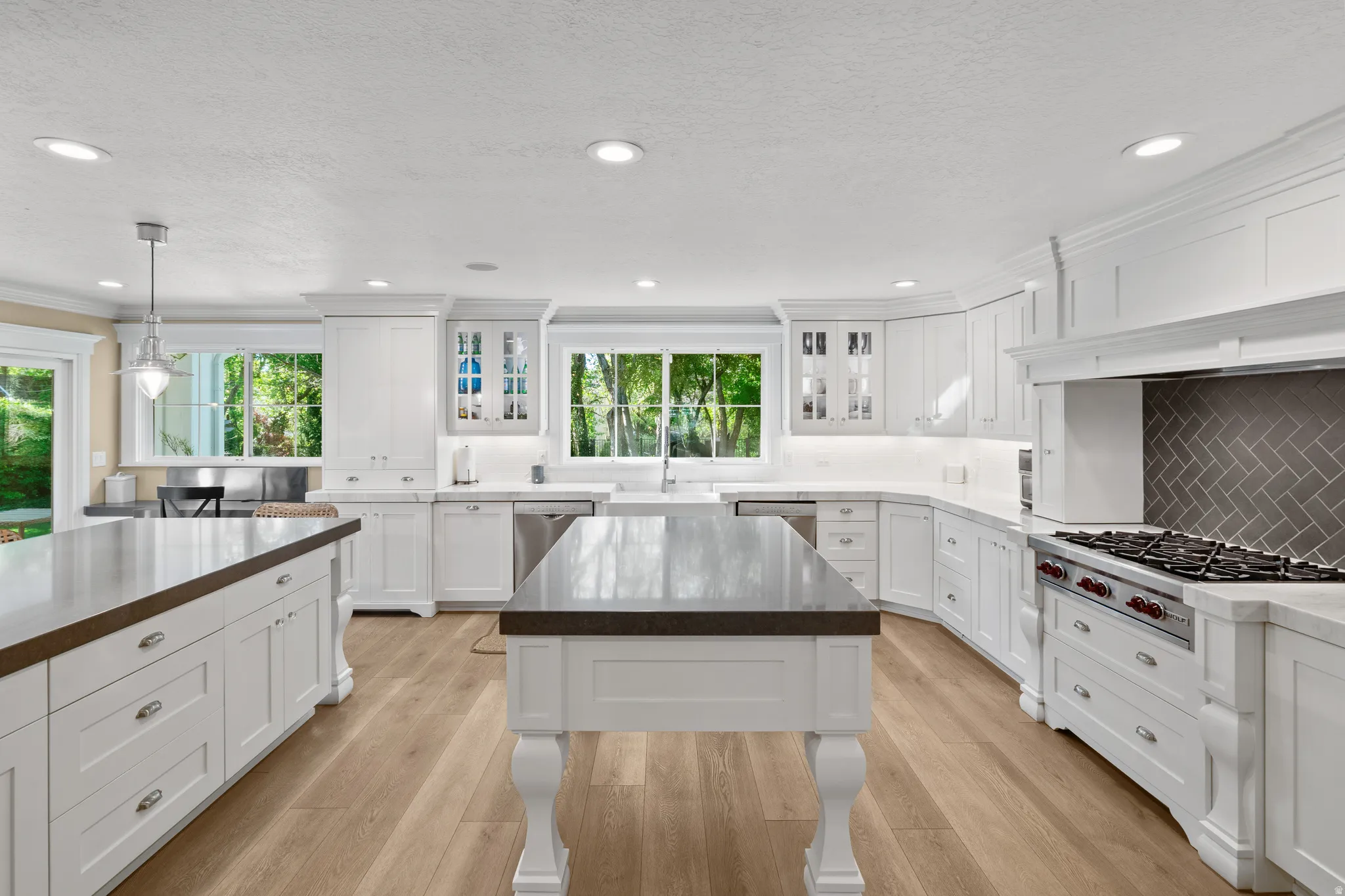 Spacious kitchen featuring two islands with dark countertops, wood-finish flooring, white cabinetry, a built-in stainless steel range, and a herringbone-patterned tile backsplash