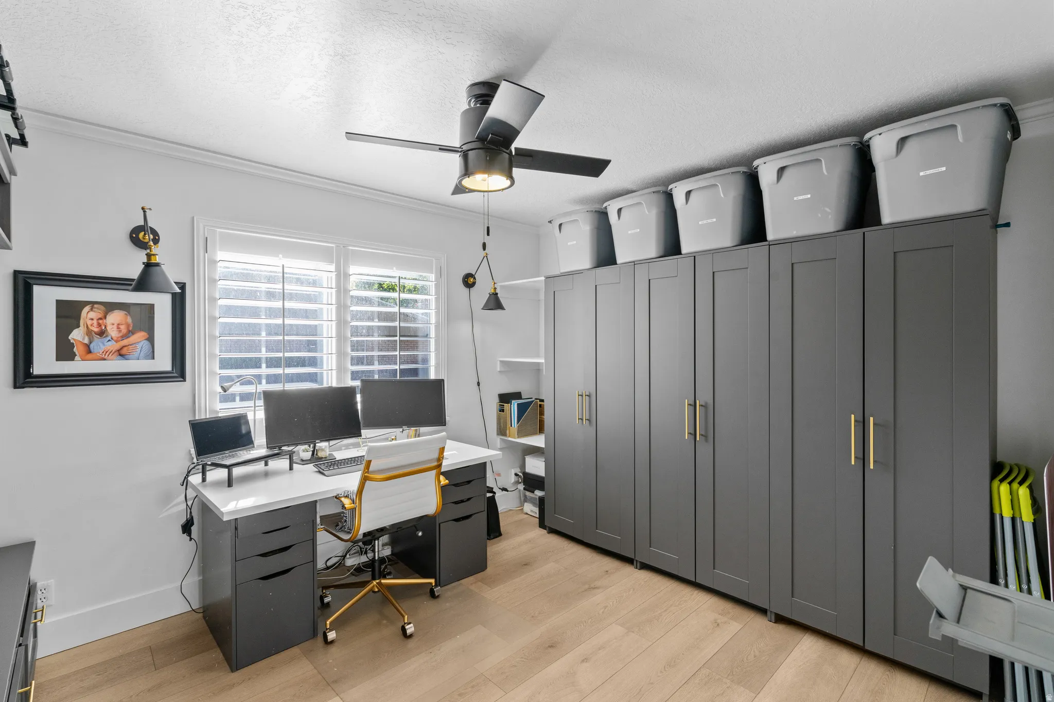 Room featuring wood-finish flooring, a ceiling fan with integrated lighting, built-in storage cabinetry with gold-tone hardware, and a window with white louvered shutters