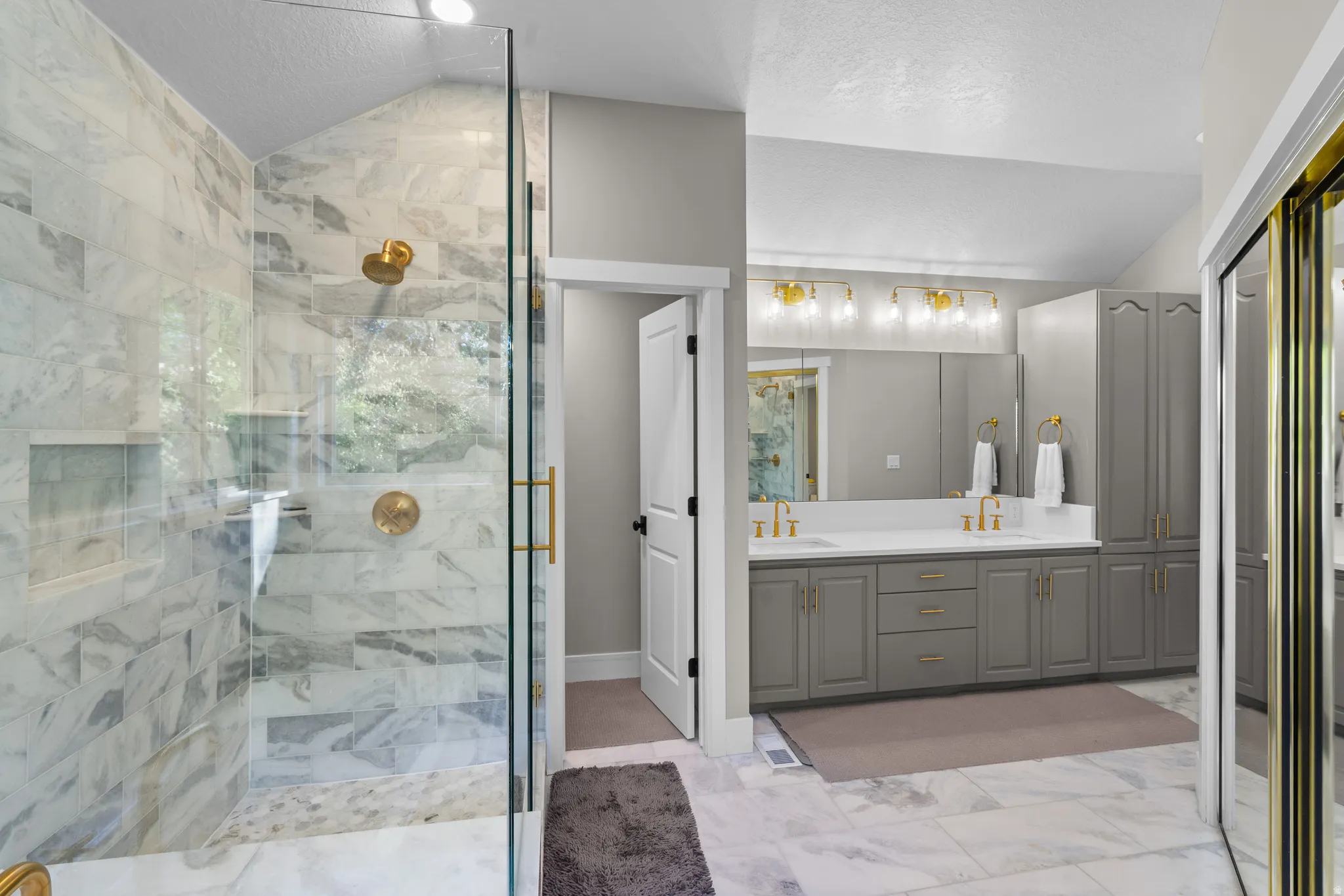 Spacious bathroom featuring a glass-enclosed shower with marble-style tile, dual vanity with white countertops, gray cabinetry, gold-finish hardware, and a vaulted ceiling