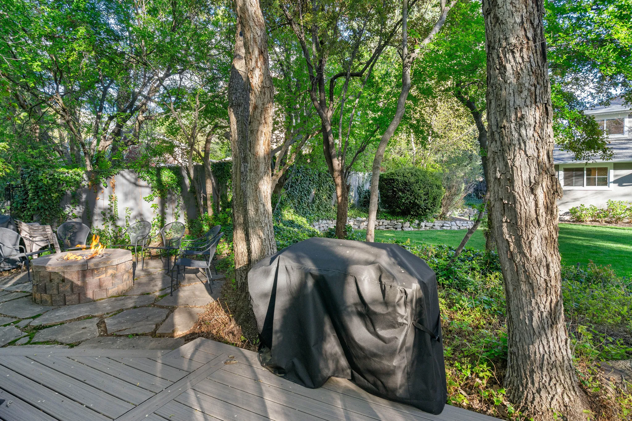 Expansive outdoor space featuring a composite deck, a stone fire pit area with flagstone pavers, mature trees, a manicured lawn, and a privacy wall