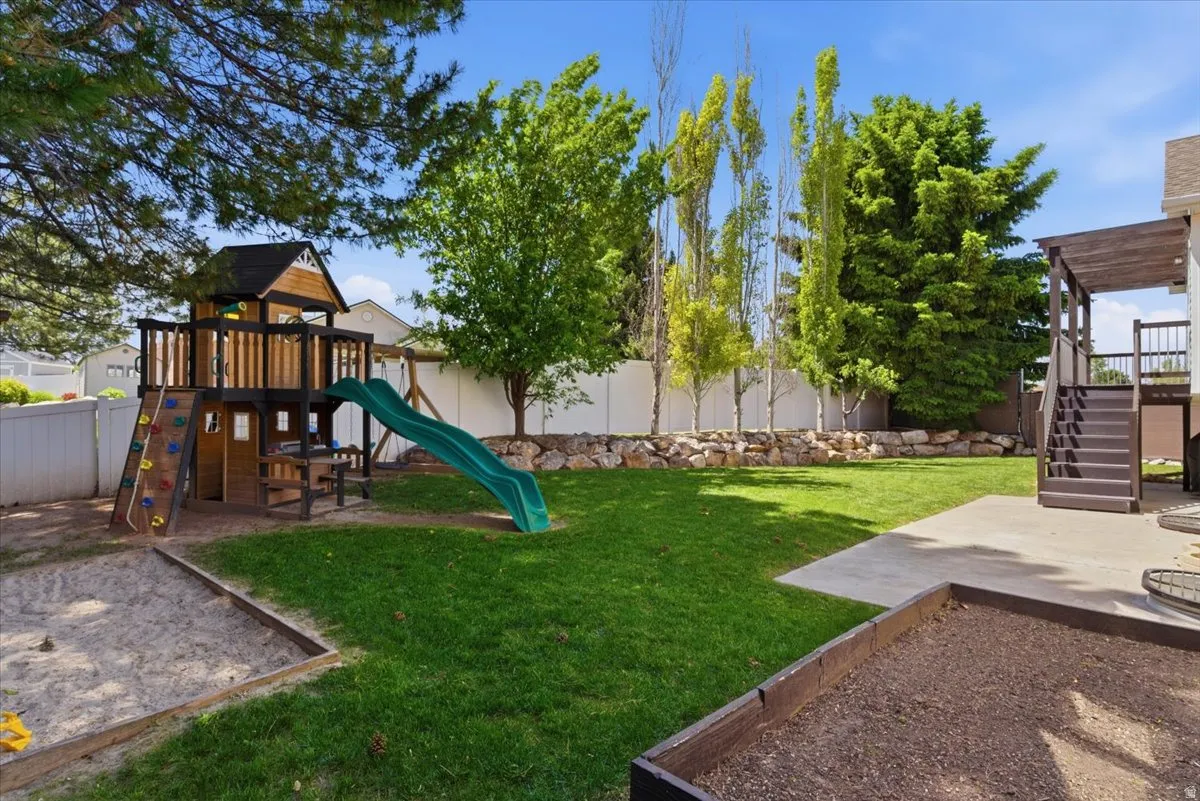 Open backyard featuring a lush lawn, mature trees, concrete patio, built-in planter box, built-in sandbox, and playground