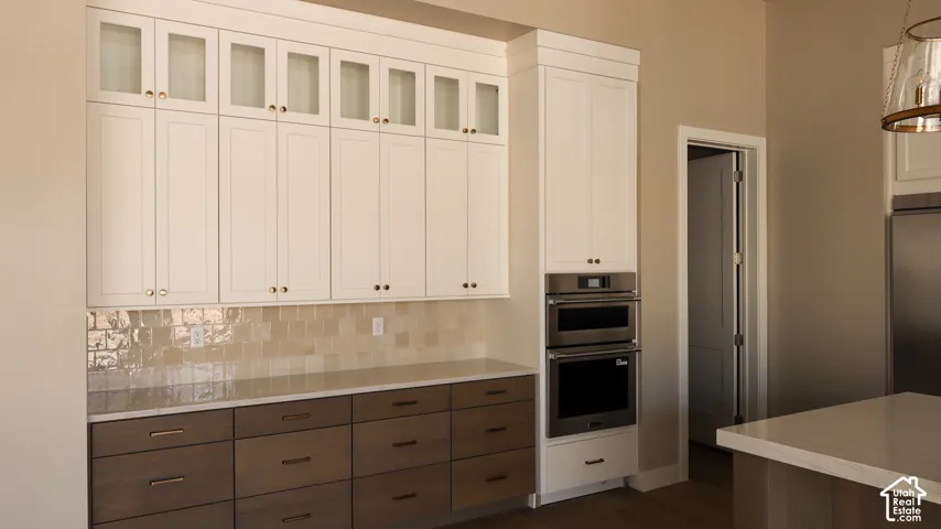Sophisticated kitchen featuring white shaker-style cabinetry with glass-front upper doors, contrasting dark wood-finish lower cabinets with multiple drawers, subway tile backsplash, stainless steel double wall ovens, integrated refrigerator, and a large c