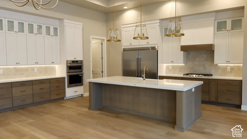 Chef's kitchen showcasing a large waterfall-edge island with quartz countertops, custom white cabinetry with glass-front uppers, stainless steel appliances including dual ovens and a gas cooktop, pendant lighting, and warm wood-finish flooring throughout