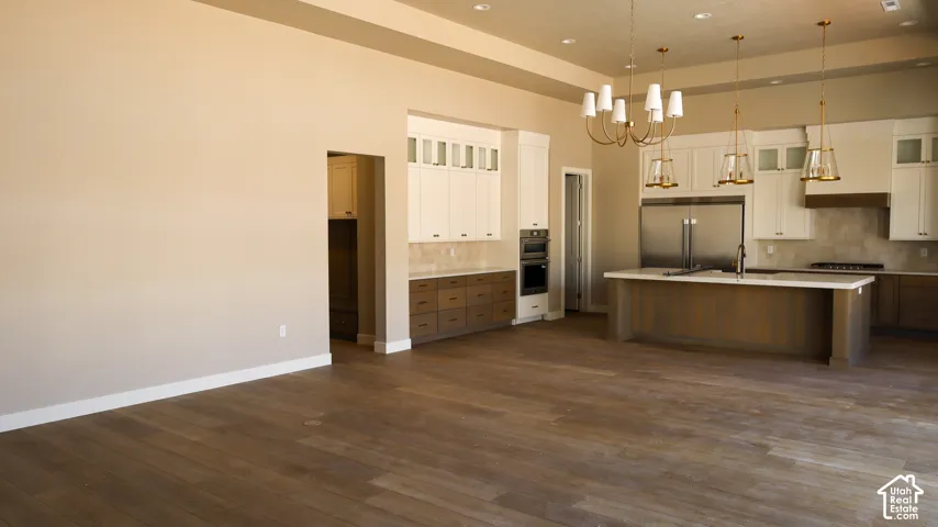 Open-concept living and kitchen area featuring dark wood-finish flooring throughout, white shaker-style cabinetry, stainless steel appliances, a large center island with seating, pendant lighting, recessed ceiling lights, and a coffered ceiling detail