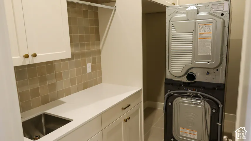 Utility room featuring stacked washer and dryer units, white cabinetry with hardware accents, neutral tile backsplash, stainless steel sink with single-basin configuration, and ample countertop workspace