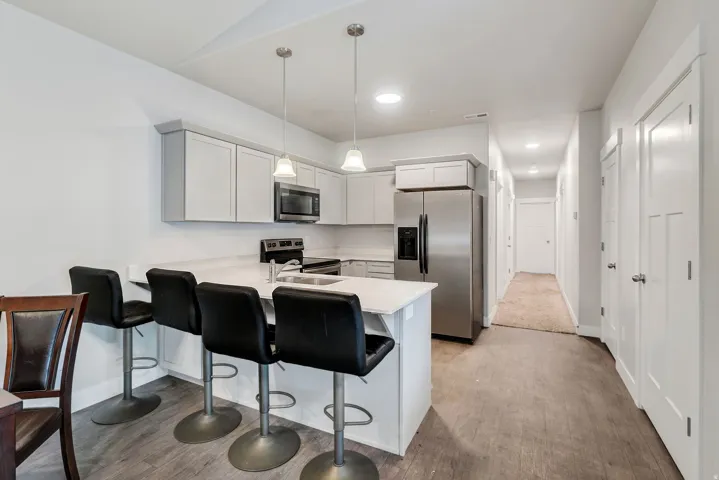 Kitchen featuring light grey cabinetry, stainless steel appliances, a breakfast bar, and wood-finish flooring extending into a hallway with recessed lighting