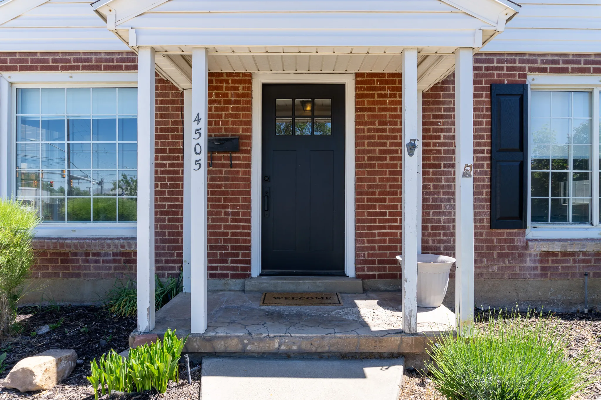 Brick facade featuring a dark paneled front door with upper glass inserts, white framed windows with gridded panes, black louvered shutters, and a covered entryway with white support columns