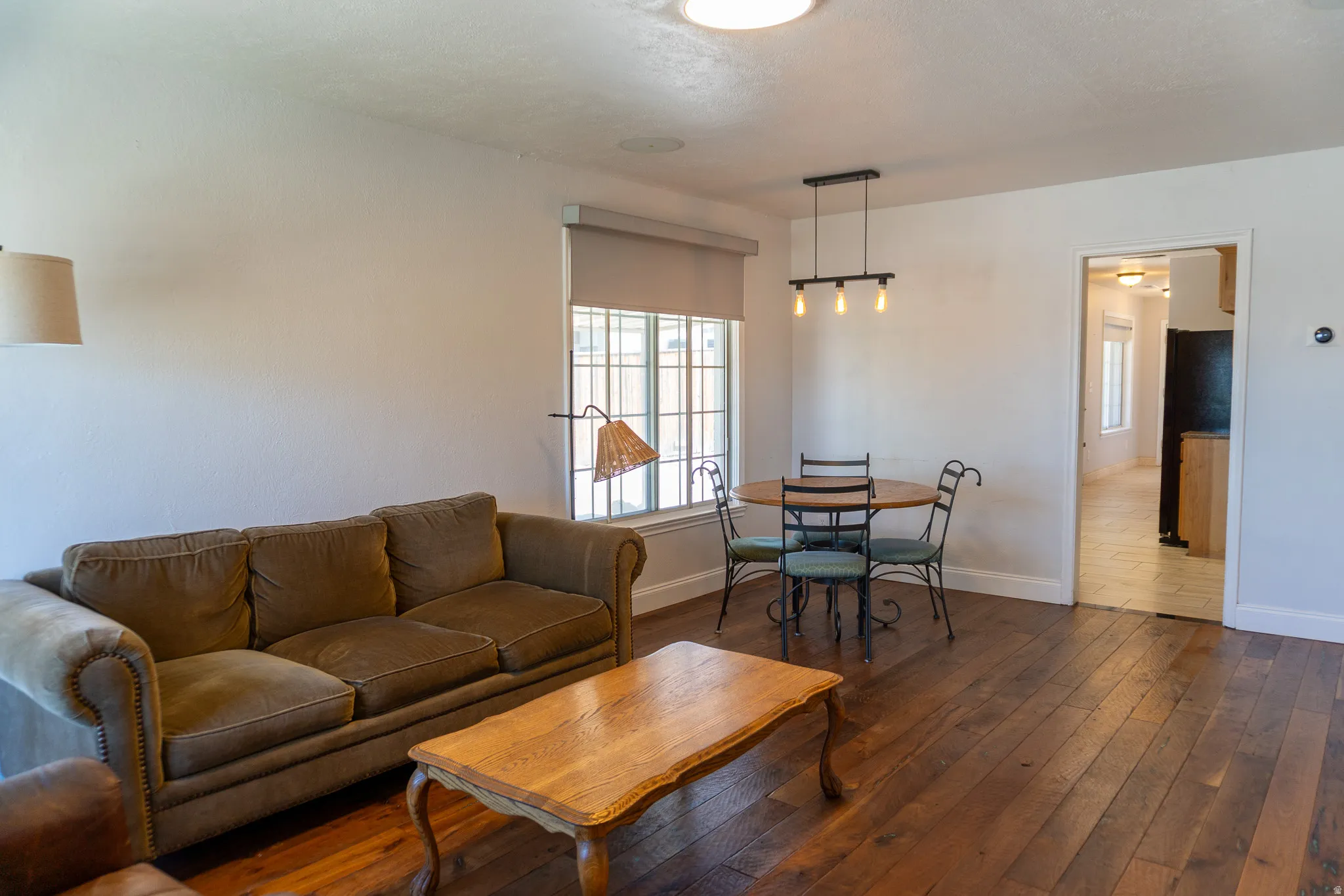 Spacious living area featuring wood-finish flooring, ample natural light from a large window, a modern linear chandelier, and an open doorway to an additional room