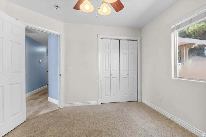 Carpeted room featuring a ceiling fan with light fixture, bifold closet doors, a window with blinds, and a doorway opening to a hallway with wood-finish flooring