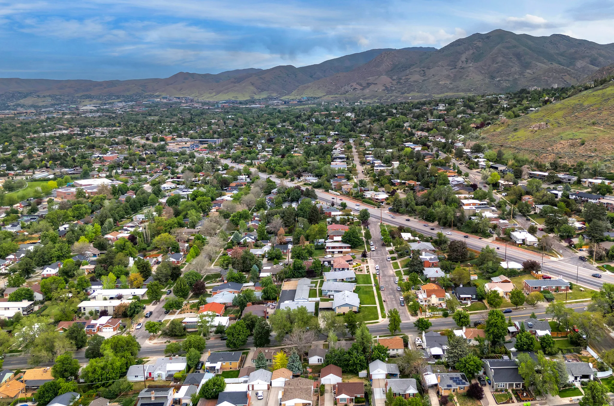 Expansive aerial view showcasing a residential area with mature tree coverage, a prominent roadway, and a distant mountain range backdrop