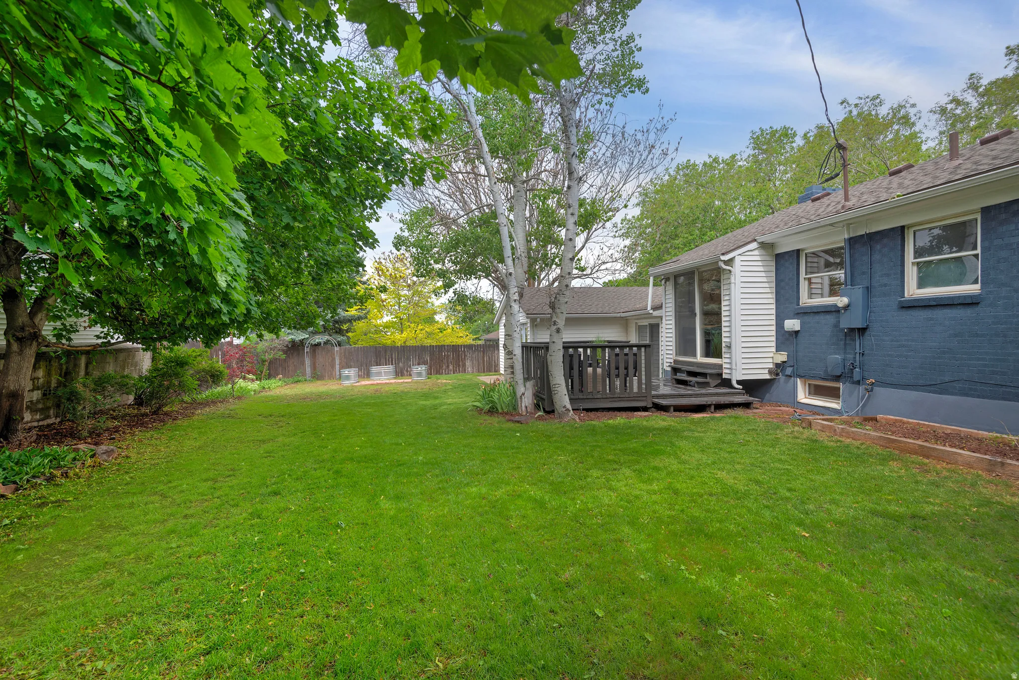 Spacious backyard featuring a large grass area, mature trees, a privacy fence, and an outdoor deck with direct access to the home