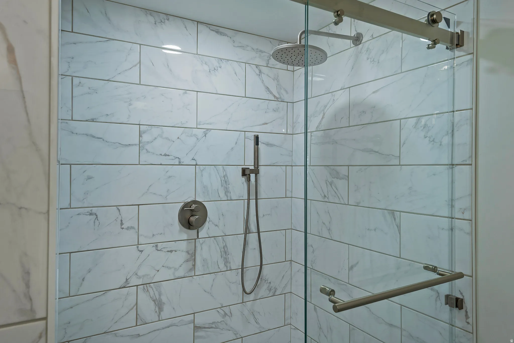 Shower enclosure featuring white marbled subway tile, a brushed nickel rainfall showerhead, a hand-held sprayer, and a frameless glass door with brushed nickel hardware