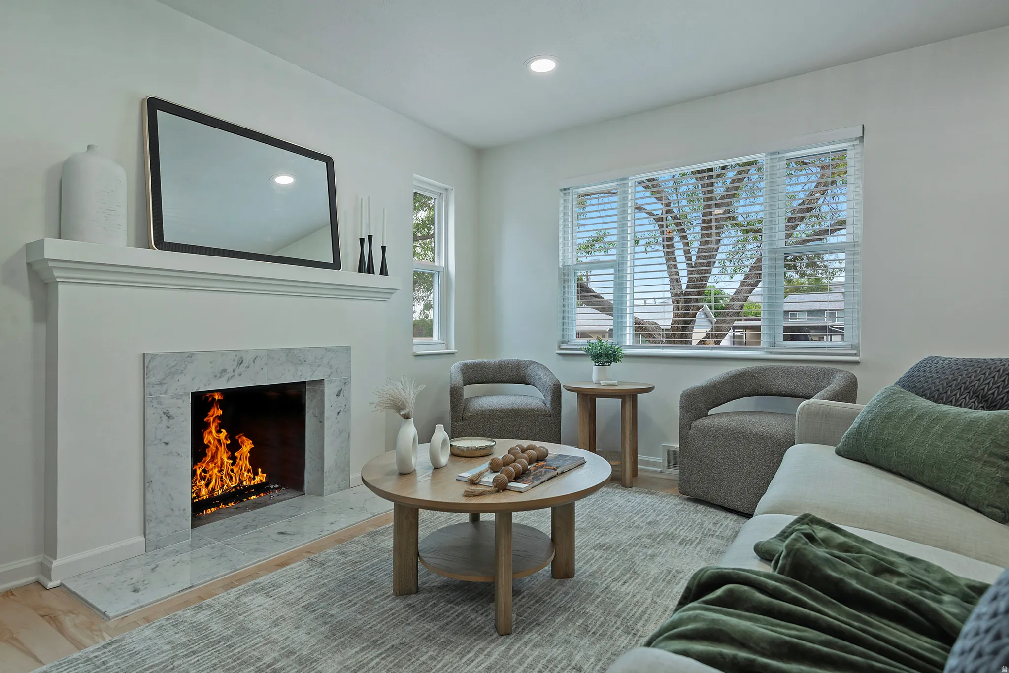 Living area featuring a white stone-surround fireplace with a classic mantel, recessed ceiling lighting, light wood-finish flooring, and a large window with blinds
