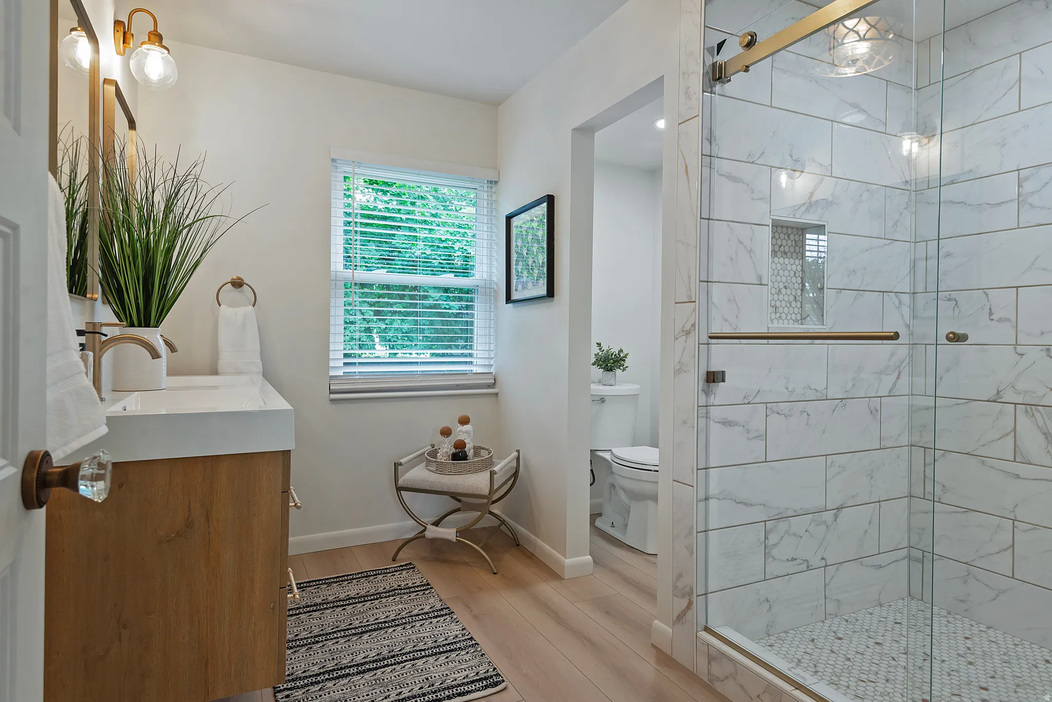 Contemporary bathroom featuring a spacious glass-enclosed shower with subway tile and hexagon tile flooring, a single vanity with a white countertop and wood-finish cabinetry, light wood-finish flooring, and a window with white blinds