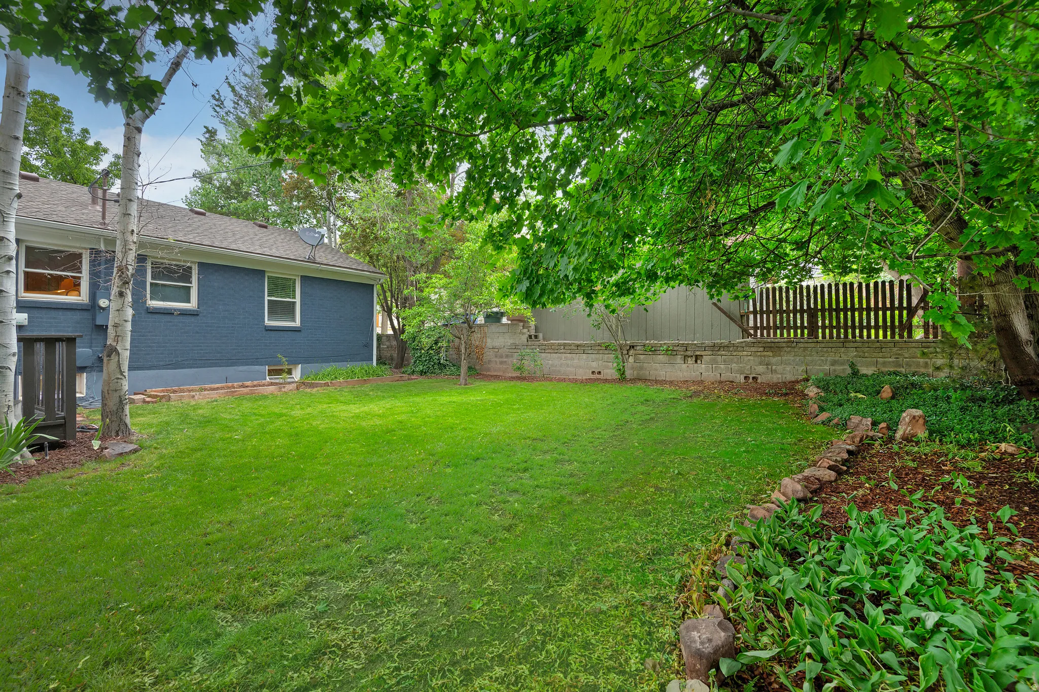 Spacious backyard featuring a lush green lawn, mature trees providing natural shade, a dark blue exterior siding on the residence, a partial privacy fence, and a stone-edged garden bed