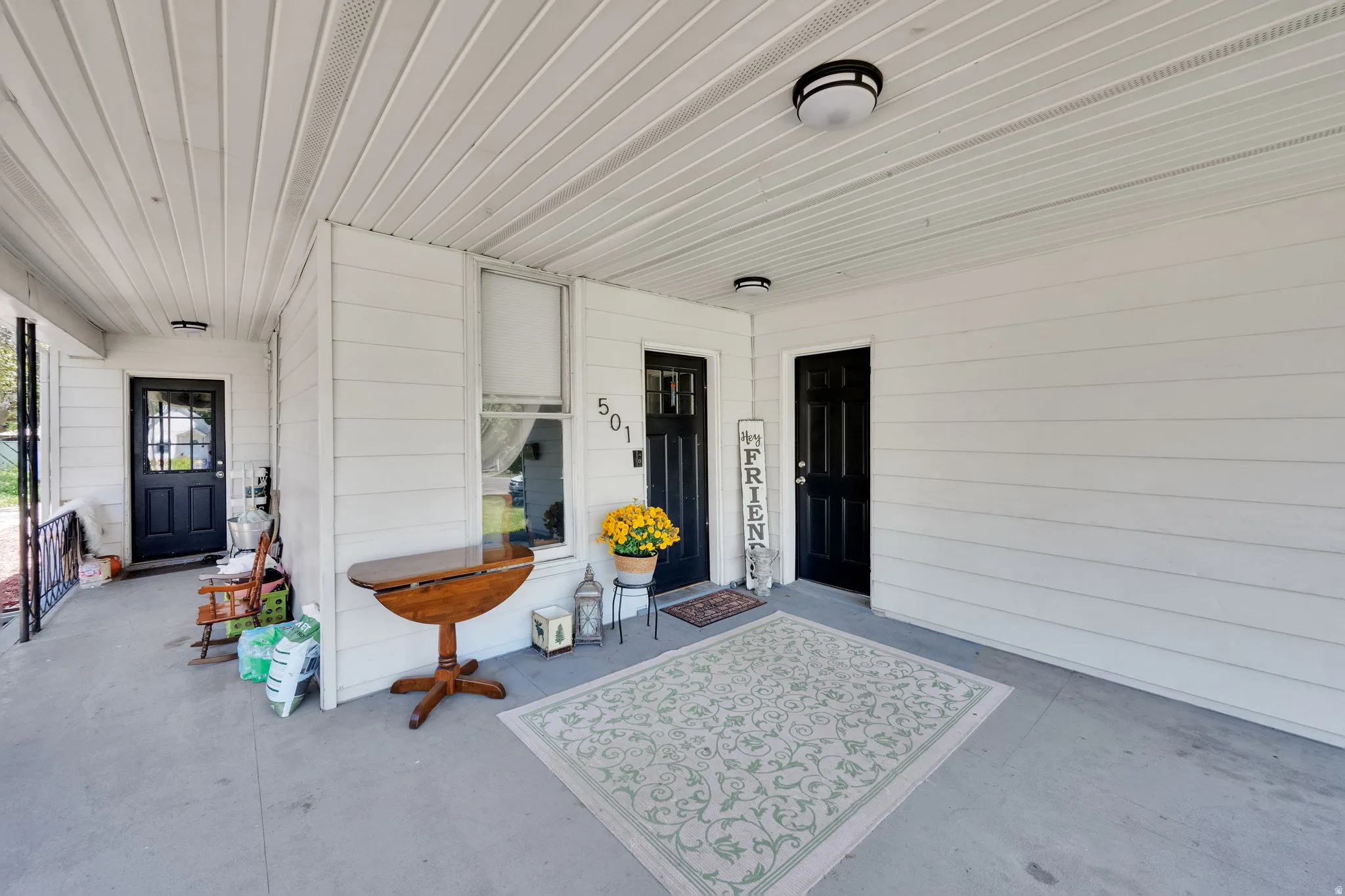 Covered front porch featuring two black paneled entry doors, white siding, a window with white trim, and overhead lighting
