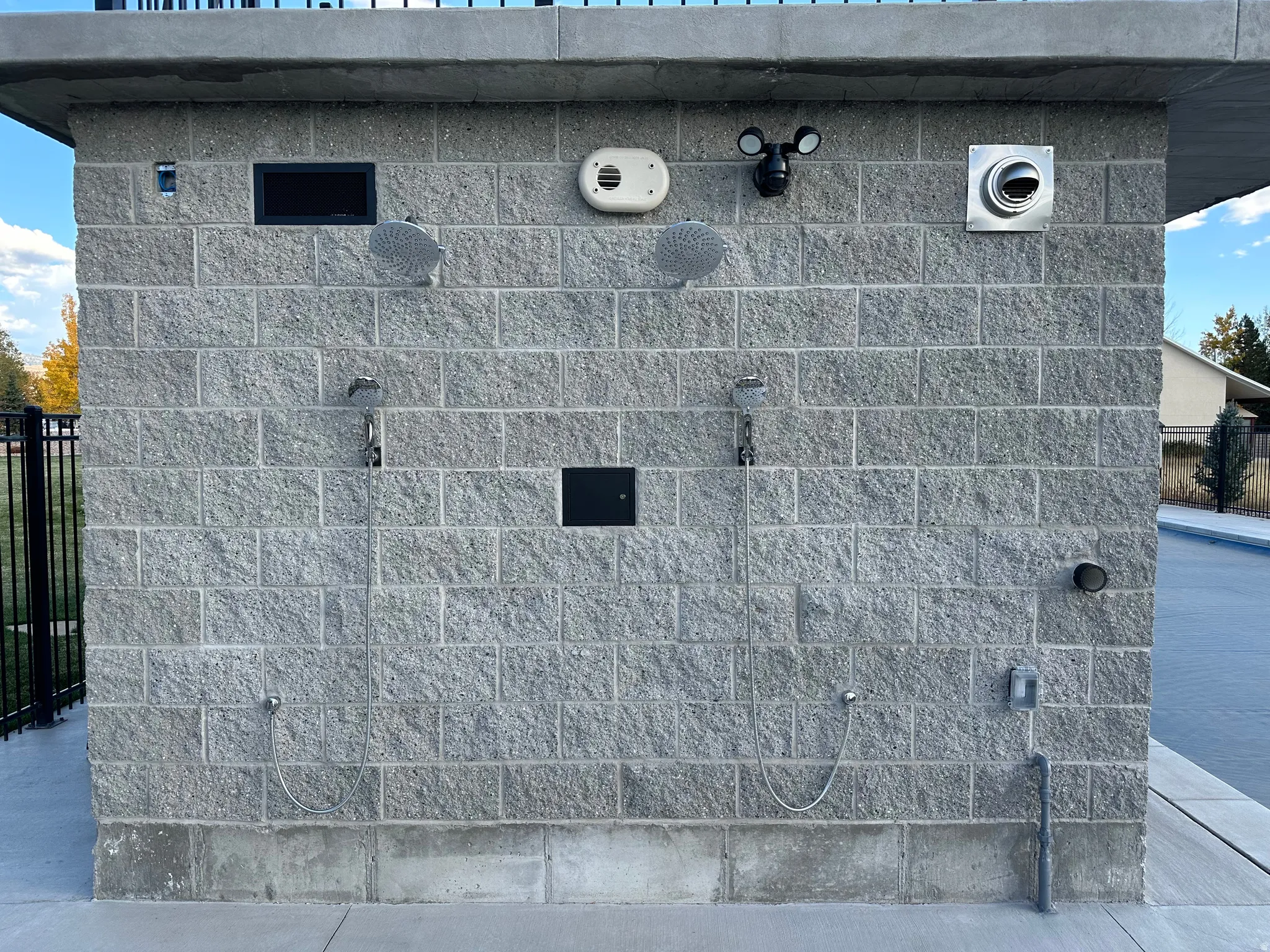 Exterior block wall structure featuring two outdoor shower fixtures with handheld sprayers, a wall-mounted anemometer, and a built-in exhaust vent