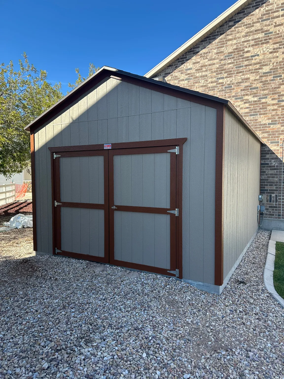 Exterior storage shed featuring a gabled roof, double entry doors with brown trim, and a gravel base