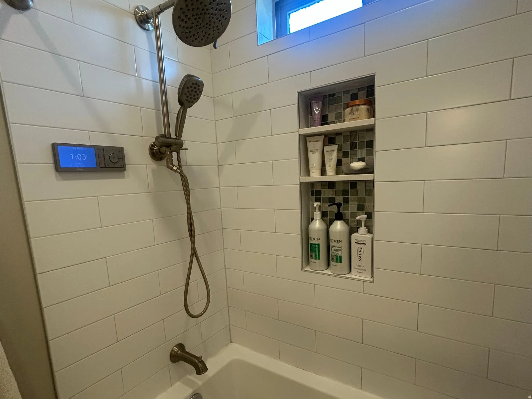 Bathtub and shower combination featuring white subway tile surround, recessed shower niche with mosaic tile accents, integrated digital shower control, and a transom window