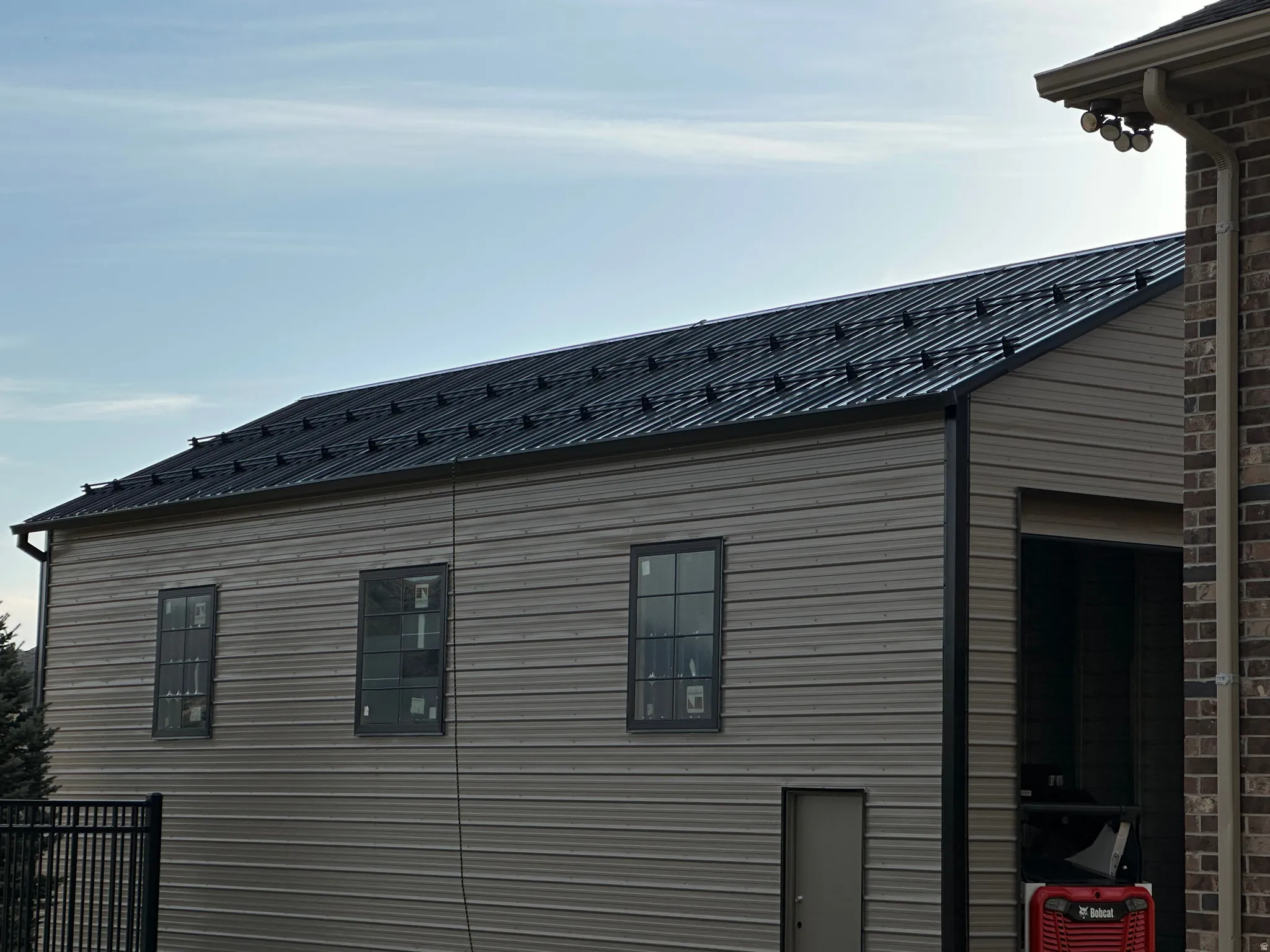 Detached outbuilding with a dark metal standing seam roof, light beige horizontal siding, and multiple dark-framed windows