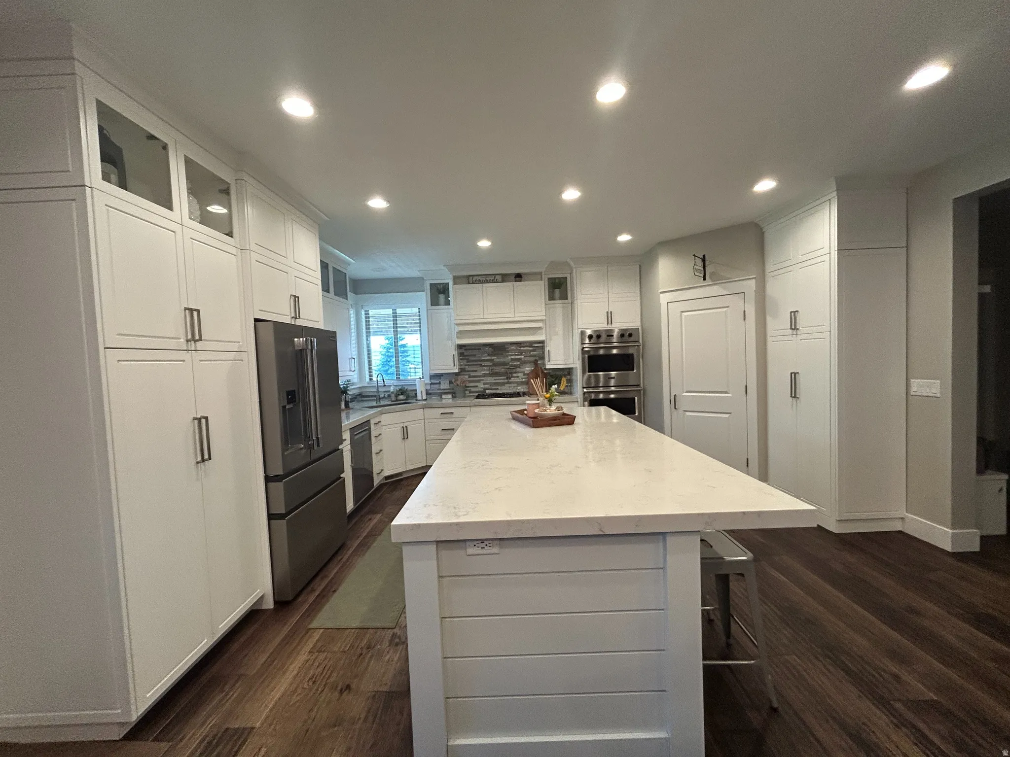 Expansive kitchen featuring a large central island with a light-toned countertop, extensive white cabinetry, wood-finish flooring, recessed lighting, and stainless steel appliances