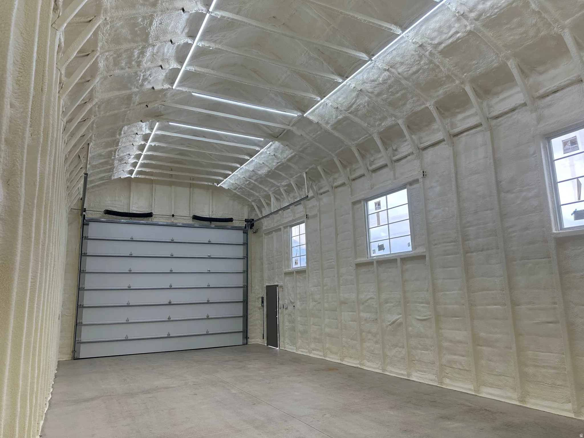 Spacious interior featuring a large overhead door, multiple windows, textured spray foam insulation on walls and ceiling, and a concrete floor