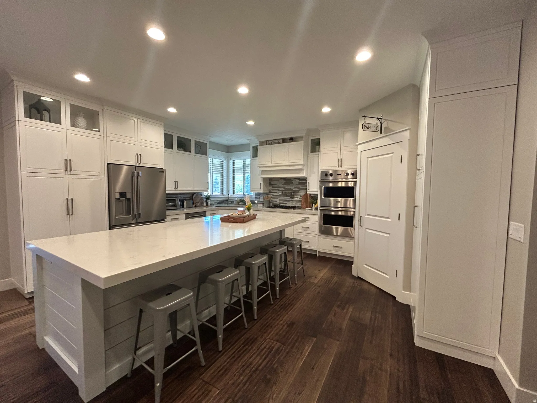 Spacious kitchen featuring a large central island with seating, ample white cabinetry, stainless steel appliances including double ovens and a refrigerator, and rich wood-finish flooring