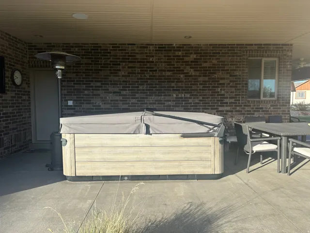 Covered patio featuring a hot tub with a light wood-finish exterior, concrete flooring, and a brick exterior wall