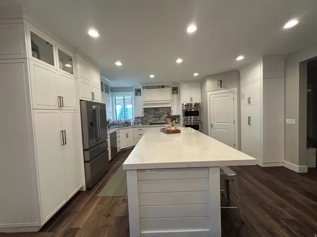 Expansive kitchen featuring a large central island with a light-toned countertop, extensive white cabinetry, wood-finish flooring, recessed lighting, and stainless steel appliances