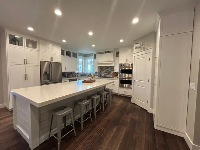 Spacious kitchen featuring a large central island with seating, ample white cabinetry, stainless steel appliances including double ovens and a refrigerator, and rich wood-finish flooring