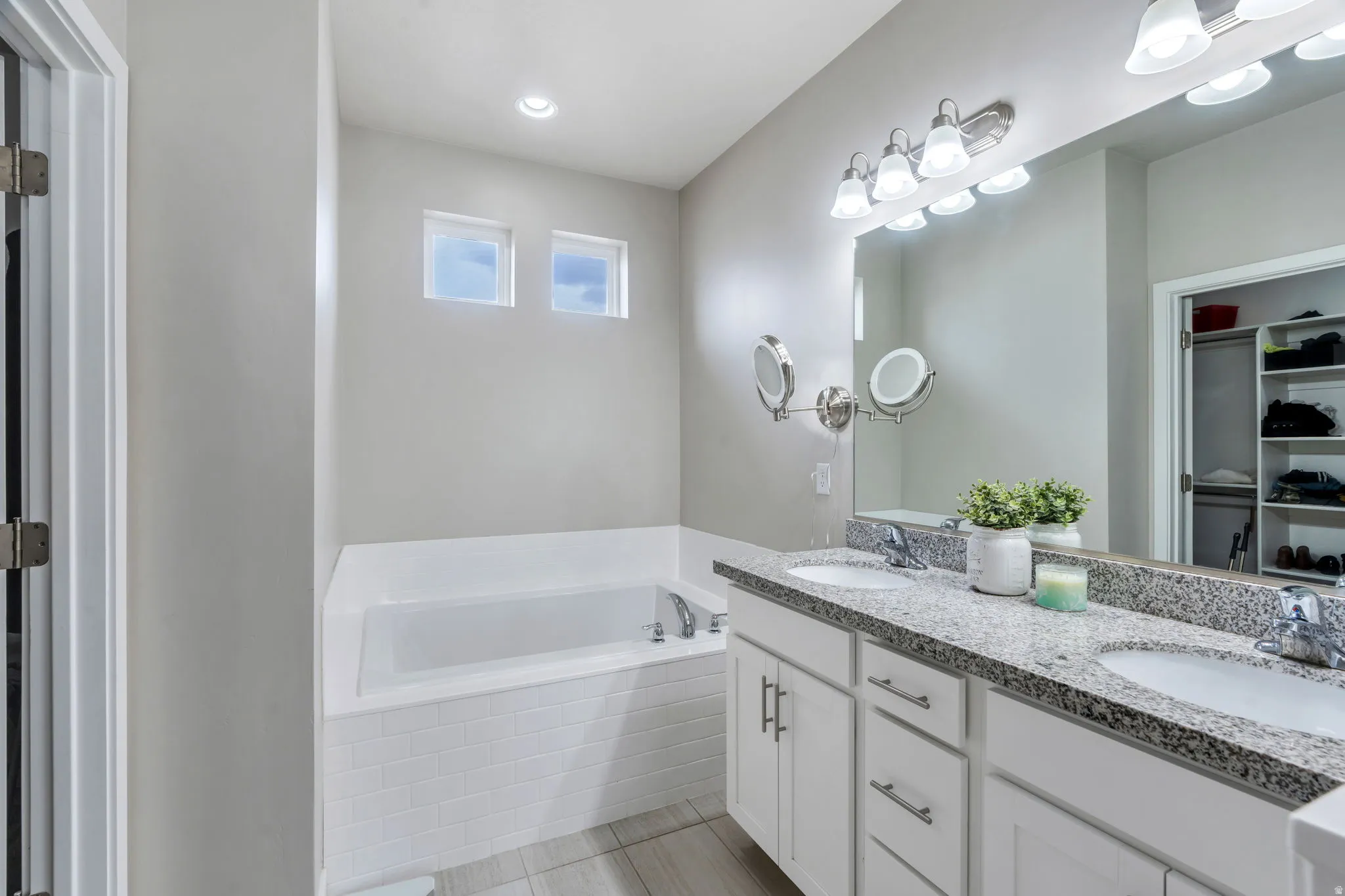 Spacious bath featuring a built-in tub with subway tile surround, two upper windows, a double vanity with granite countertop, and integrated lighting