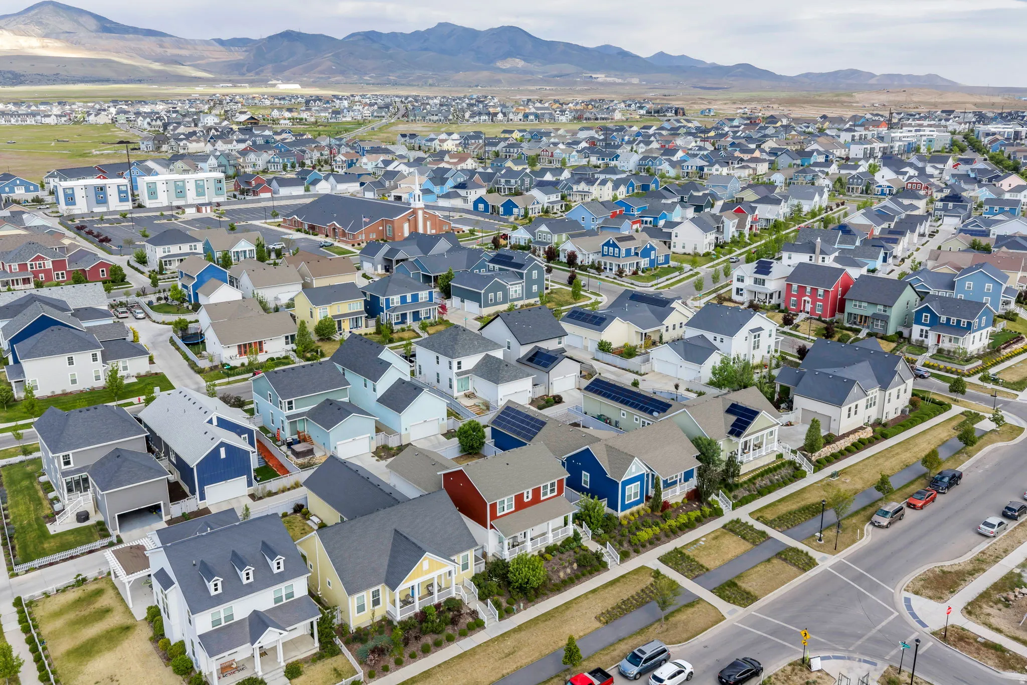 Expansive residential development featuring a variety of architectural styles, individual garages, solar panels visible on multiple rooftops, numerous street trees, and mountainous scenery