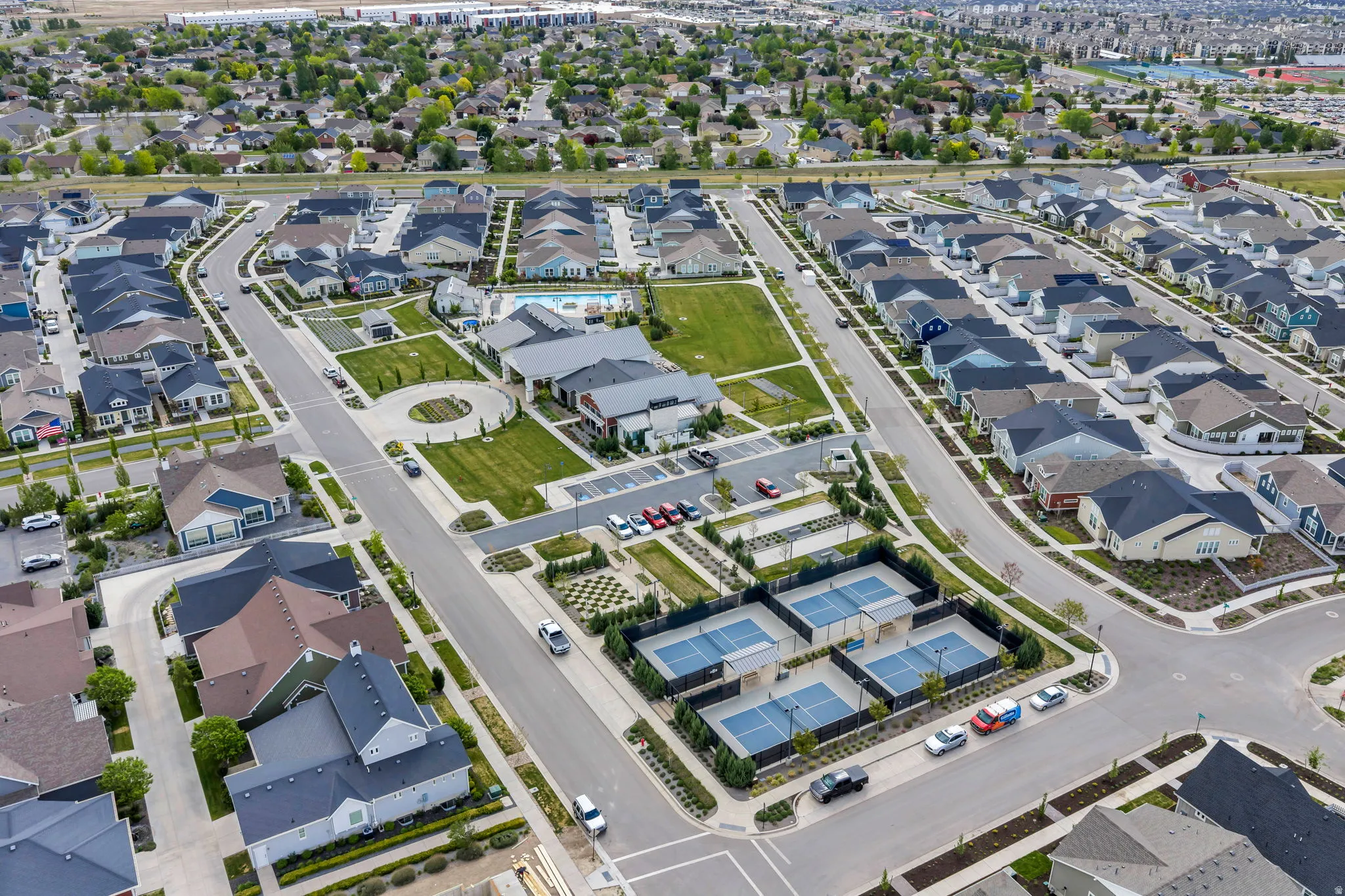 Aerial view of a residential community featuring multiple modern homes, a central green space, a community pool, and several sports courts