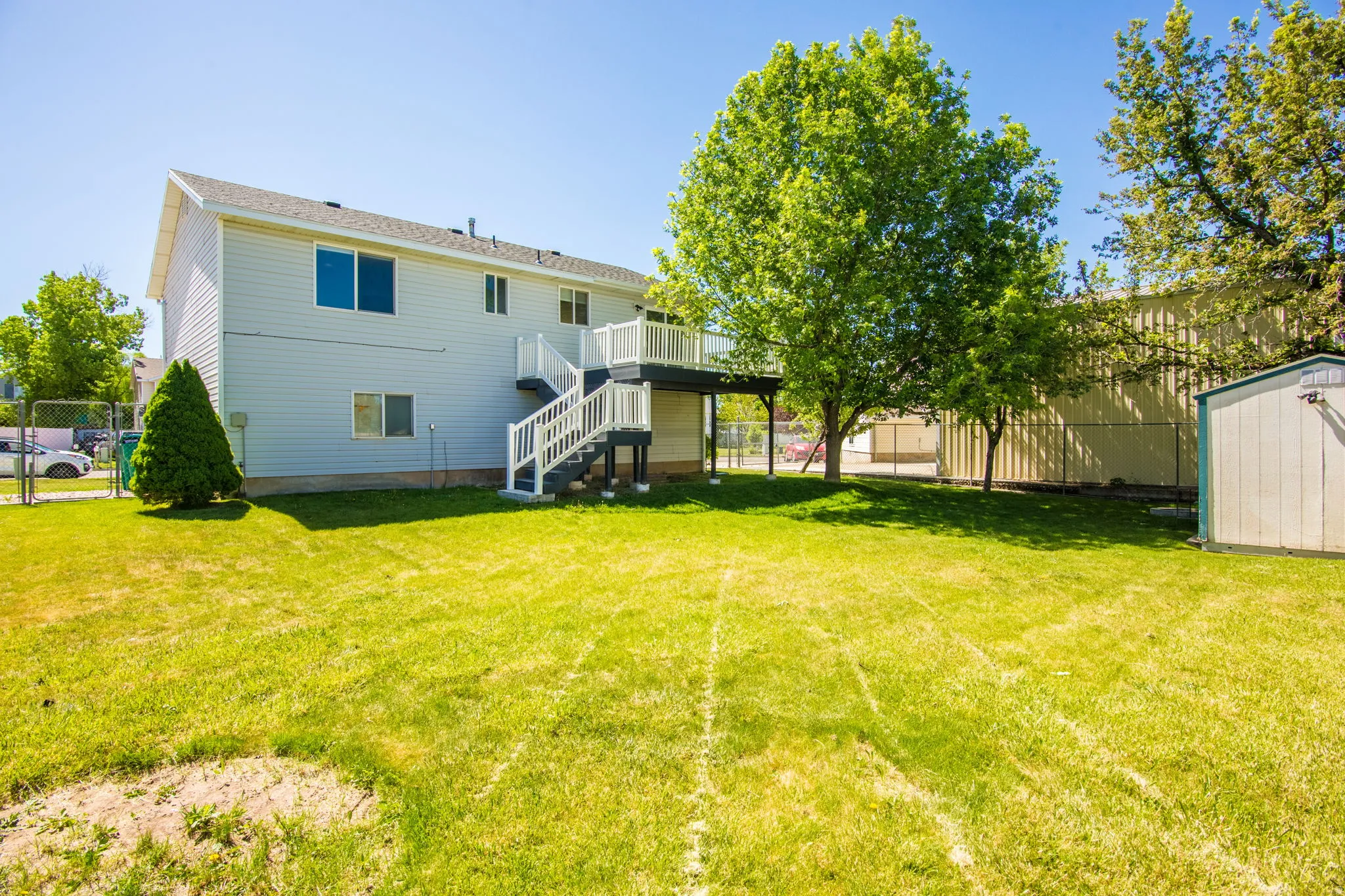 Spacious backyard featuring a two-story residence with light-colored siding, a second-story deck with white railings, and mature trees providing shade