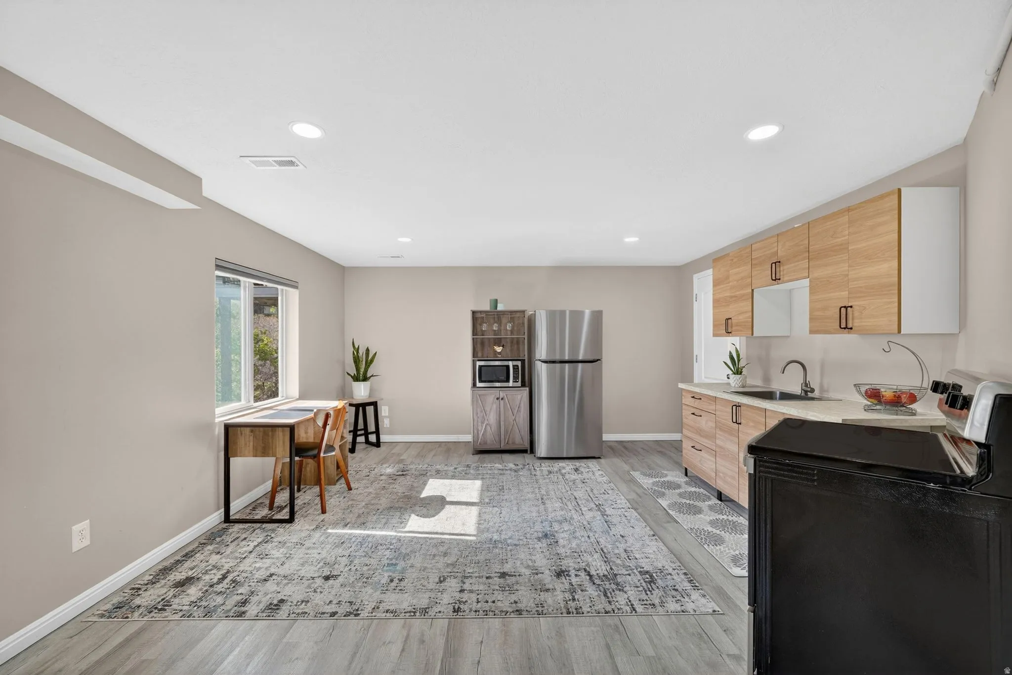 Spacious open-concept room featuring light wood-finish flooring, a contemporary kitchen area with wood-tone cabinetry, stainless steel sink, and light countertops