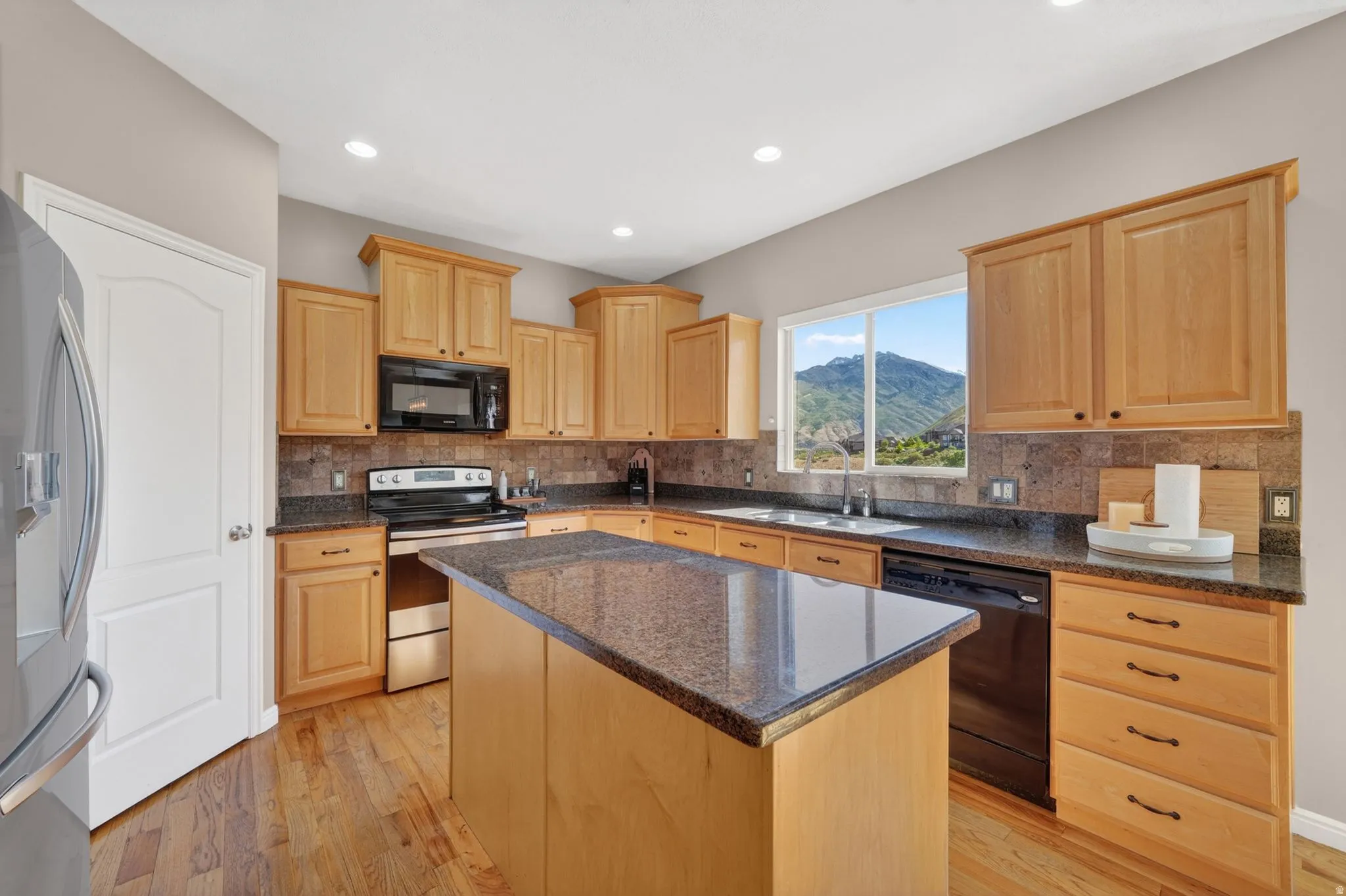 Kitchen featuring wood-finish flooring, a central island with a stone-finish countertop, wood cabinetry, built-in black and stainless steel appliances, and a window framing a mountain backdrop
